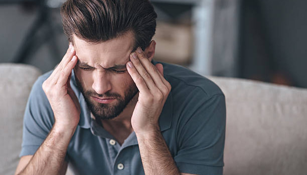 Frustrated handsome young man touching his head and keeping eyes closed while sitting on the couch at home