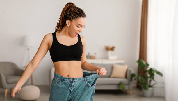 Excited Slim Black Female After Great Weight Loss Showing Result Of Slimming Wearing Old Big Jeans Standing Indoors. Successful Dieting And Slimming Concept