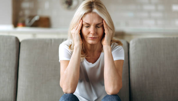 Middle aged blonde woman sits on couch at living room holding her head with her hands, feels unhappy because of headache, personal troubles, illness or bad news, she need psychological or medical support